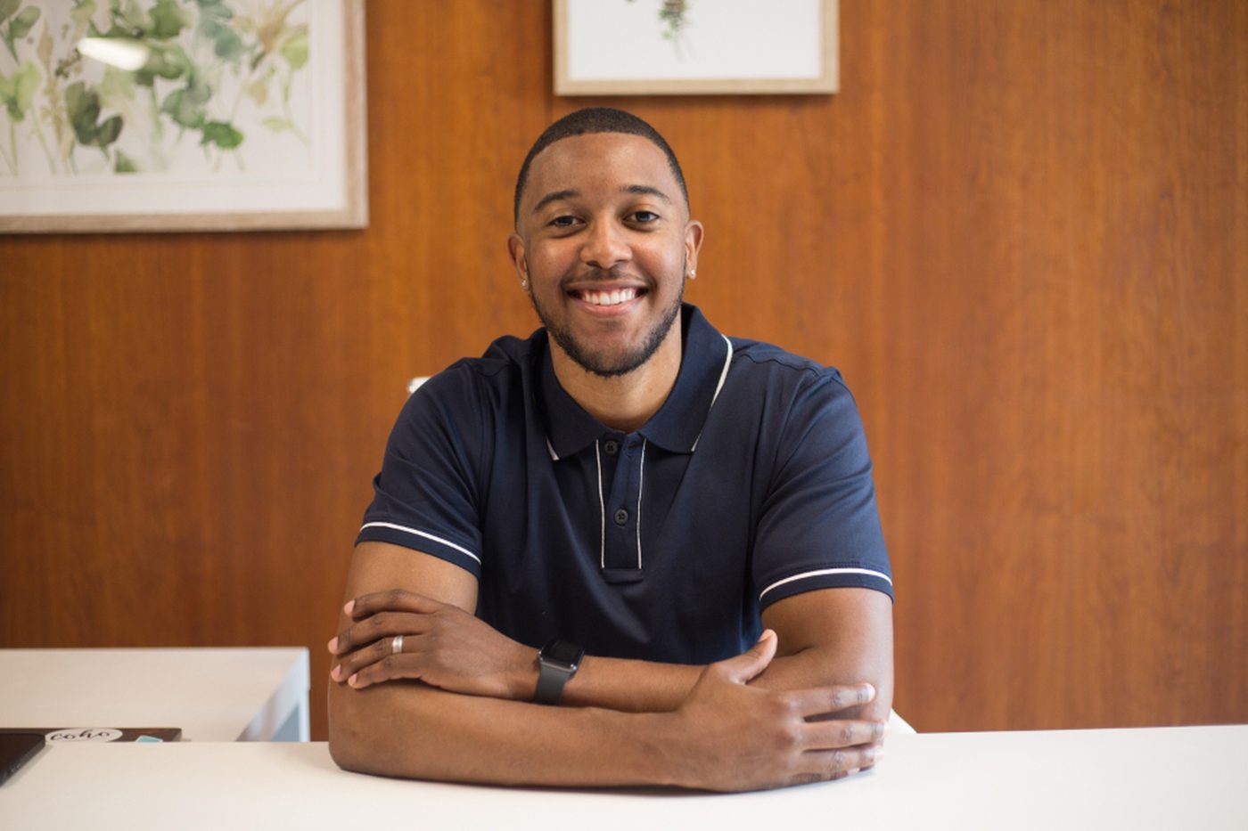 A smiling man in a navy polo shirt sitting at a desk